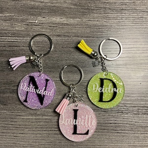 May include: Three glittery round keyrings with silver key rings and tassel accents. Each keyring features a letter and a name: "N" with "Natividad", "L" with "Laurelle", and "D" with "Deidra".