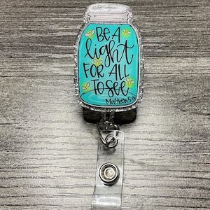 May include: A badge reel featuring a teal jar design with the words "Be a light for all to see" in black script. The jar has a silver glitter border and the text "Matthew 5:16". The reel has a clear plastic clip.