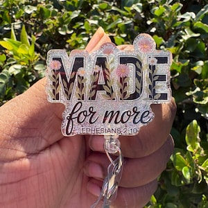 May include: A clear badge reel with the words "MADE for more" in black and silver glitter. The letters are decorated with pink and white flowers. The text "Ephesians 2:10" is written below the words "for more".