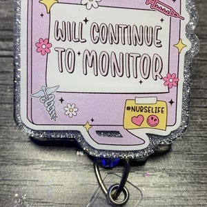 May include: A badge reel with a light purple background and a silver glitter border. The badge reel has the text "WILL CONTINUE TO MONITOR" with a caduceus, flowers, stars, and a computer graphic with the hashtag "#NURSELIFE".