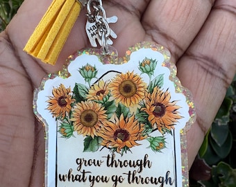 May include: A clear acrylic keyring featuring a sunflower design and the text "grow through what you go through." The keyring has a yellow tassel and silver-coloured hardware. The design features orange and yellow sunflowers with green leaves.