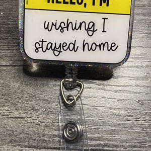 May include: A rectangular badge reel with a yellow top and a white bottom. The text "HELLO, I'M" is above "wishing I stayed home" in black script. The badge has a clear plastic strap and a silver metal clip.