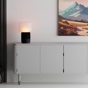 Può includere: Una moderna lampada da tavolo in bianco e nero con un paralume in vetro smerigliato, che emette un bagliore caldo. La lampada è su un mobile grigio chiaro con un dipinto di un paesaggio montano sopra. Una borsa nera è appoggiata sul pavimento.