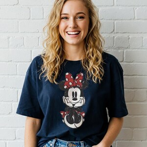 May include: A navy blue t-shirt featuring a distressed graphic of Minnie Mouse with a red polka dot bow. The cartoon character is centered on the shirt. The person is wearing blue jeans and is smiling.