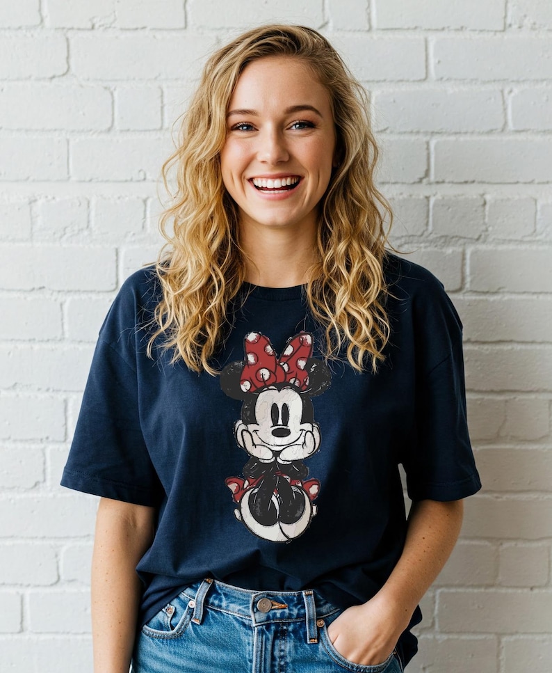 May include: A navy blue t-shirt featuring a distressed graphic of Minnie Mouse with a red polka dot bow. The cartoon character is centered on the shirt. The person is wearing blue jeans and is smiling.