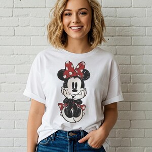 May include: White t-shirt featuring a distressed graphic of Minnie Mouse with a red polka dot bow. The cartoon character is in a seated pose. The shirt has rolled-up sleeves and is paired with blue jeans.
