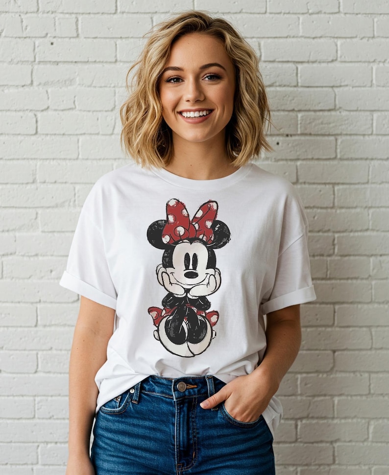 May include: White t-shirt featuring a distressed graphic of Minnie Mouse with a red polka dot bow. The cartoon character is in a seated pose. The shirt has rolled-up sleeves and is paired with blue jeans.