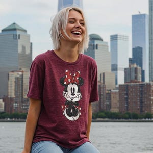 May include: Maroon t-shirt featuring a Minnie Mouse graphic. The graphic shows Minnie Mouse with a red polka dot bow. The shirt is made of a soft material and is paired with blue jeans. The background features a cityscape with tall buildings and a body of water.