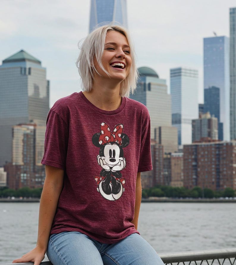 May include: Maroon t-shirt featuring a Minnie Mouse graphic. The graphic shows Minnie Mouse with a red polka dot bow. The shirt is made of a soft material and is paired with blue jeans. The background features a cityscape with tall buildings and a body of water.