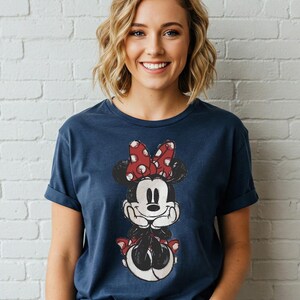 May include: Navy blue t-shirt featuring a distressed graphic of Minnie Mouse with a red polka dot bow. The shirt has rolled-up sleeves and is worn by a person in front of a white brick wall.