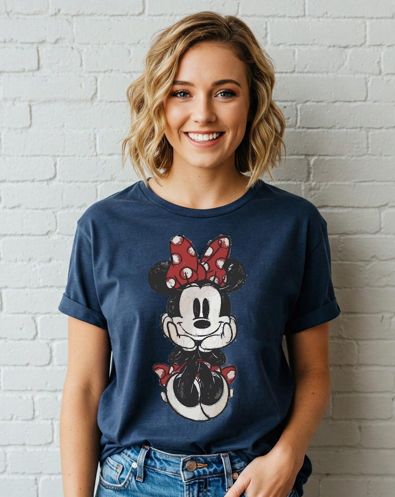 May include: Navy blue t-shirt featuring a distressed graphic of Minnie Mouse with a red polka dot bow. The shirt has rolled-up sleeves and is worn by a person in front of a white brick wall.