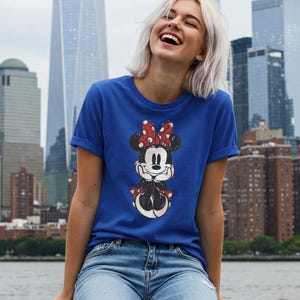 May include: Royal blue t-shirt featuring a Minnie Mouse graphic. The graphic shows Minnie Mouse in a classic pose with a red bow and polka dots. The shirt has short sleeves and is paired with blue jeans.