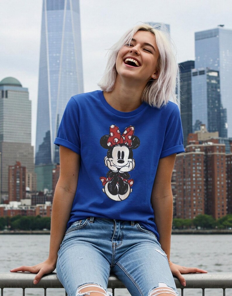 May include: Royal blue t-shirt featuring a Minnie Mouse graphic. The graphic shows Minnie Mouse in a classic pose with a red bow and polka dots. The shirt has short sleeves and is paired with blue jeans.