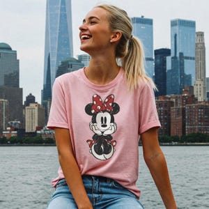 May include: A light pink t-shirt featuring a Minnie Mouse graphic. The graphic shows Minnie Mouse with a red bow with white polka dots. The t-shirt has short sleeves and a crew neck.
