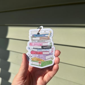 May include: A die-cut sticker featuring a stack of books with titles including The Tortured Poets Department, Midnights, evermore, folklore, Lover, reputation, 1989, RED, Speak Now, FEARLESS, and Taylor Swift, decorated with floral and snake illustrations.