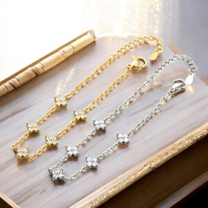 Może przedstawiać: Dwa delikatne bransoletki z łańcuszkowym wzorem i małymi kwiatowymi zawieszkami ozdobionymi przezroczystymi cyrkoniami. Jeden bransoletka jest złota, a drugi srebrny.