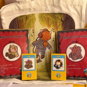 Könnte beinhalten: Eine Stofftasche mit einer Cartoon-Figur mit Kopfhörern im Wald, zwei rote Kreuzstich-Sets mit Bärenmotiven und zwei Emaille-Anstecknadeln auf gelben Karten. Die Sets enthalten Stoff, Vorlage, Anleitung, Garn und einen 7,6 cm Stickrahmen.
