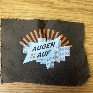 Könnte beinhalten: Ein dunkelbrauner Stofffleck mit den Worten "AUGEN AUF" in Weiß, überlagert von einer weißen geometrischen Form. Der Flicken hat orangefarbene Strahlenlinien und zusätzlichen Text in Weiß. Der Stoff hat einen rohen, unfertigen Rand.