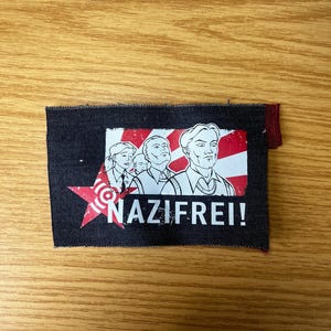 Könnte beinhalten: Ein rechteckiger Aufnäher mit dem Wort "NAZIFREI!" in weißen Großbuchstaben. Der Aufnäher zeigt eine rot-weiße Grafik eines Sterns und einer Gruppe von Personen. Der Aufnäher befindet sich auf einem dunkelblauen Hintergrund.