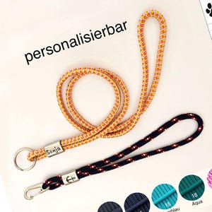 Könnte beinhalten: Zwei Schlüsselbänder aus geflochtenem Seil. Eines ist orange-gelb mit einem silbernen Metallanhänger, der den Namen Sonja trägt, und einem Ring. Das andere ist dunkelblau mit weißen und roten Sprenkeln, einem Anker-Metallanhänger und Karabiner.