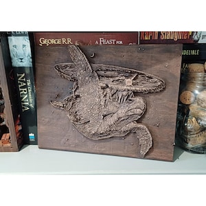 May include: A detailed, bronze-colored wooden carving of a turtle carrying a flat disc on its back. The carving is mounted on a square wooden plaque. The image is of a fantasy theme, with books in the background.