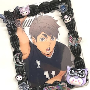 Puede incluir: Un marco de fotos decorativo con una imagen de un personaje de anime. El marco negro está adornado con adornos de mariposas, flores y personajes de dibujos animados. La imagen interior muestra un personaje de anime masculino con una camisa azul oscuro con el número 11.