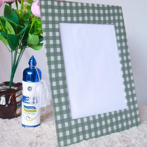 May include: A green and white gingham patterned picture frame. The frame is leaning against a white surface. A blue bottle with a white tassel and a bouquet of pink flowers are to the left of the frame.