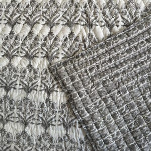Könnte beinhalten: Eine weiße und graue Steppdecke mit floralem Muster. Die Steppdecke zeigt ein sich wiederholendes Design aus stilisierten Blumen und Blättern in Grau auf weißem Hintergrund. Die Steppdecke ist mit einem Rautenmuster gesteppt.