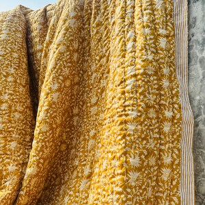 Pode incluir: Uma manta acolchoada amarelo-mostarda com um padrão floral branco repetido por todo o tecido. O material apresenta uma textura pespontada visível e é rematado com uma borda de riscas verticais em tons de amarelo-mostarda e branco.