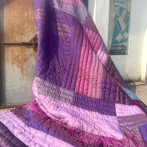 Könnte beinhalten: Eine große, gesteppte Decke in Lila- und Rosatönen. Die Decke hat ein Patchwork-Design mit unterschiedlichen Texturen und Farbtönen, wodurch ein optisch ansprechendes Muster entsteht. Der Stoff scheint weich zu sein und möglicherweise aus Seide oder Satin.