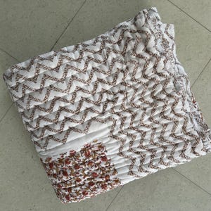 Könnte beinhalten: Weiß-braun gemusterte Steppdecke mit Zick-Zack-Design. Die Steppdecke hat einen floralen Rand in Rot-, Braun- und Goldtönen. Der Stoff ist gesteppt und erzeugt eine strukturierte Oberfläche. Dieser Bettwäscheartikel eignet sich für ein Bett oder als dekorativer Überwurf.