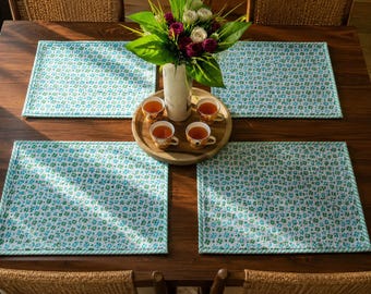 Puede incluir: Cuatro manteles individuales rectangulares con fondo blanco y un pequeño estampado floral azul y verde, con un borde a rayas azul claro y blanco. Una bandeja redonda de madera contiene cuatro tazas de té pequeñas y un florero con flores rosas y blancas.