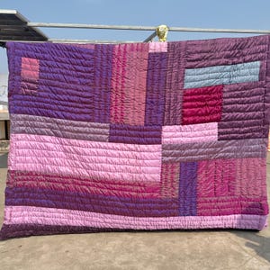 Op de afbeelding: Een gewatteerde deken in paarse en roze tinten, met een patchwork ontwerp. De deken is gemaakt van een glanzende, gewatteerde stof. Het ontwerp bevat rechthoekige en vierkante stukken in verschillende tinten paars en roze.