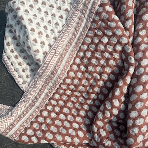 Può includere: Una coperta trapuntata piegata con un motivo floreale bianco e marrone. La coperta ha un bordo marrone con un disegno geometrico. L'altro lato della coperta ha uno sfondo marrone con un motivo a nuvola bianca.