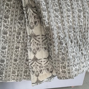 Könnte beinhalten: Eine grau-weiß gemusterte Steppdecke. Die Steppdecke hat ein sich wiederholendes Muster aus Blumen und Rauten. Die Steppdecke besteht aus einem weichen, gesteppten Material. Die Steppdecke hat eine neutrale Farbe und würde jede Schlafzimmerdekoration ergänzen.