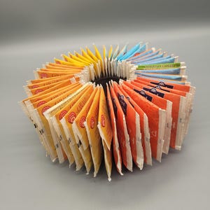 Pode incluir: Um arranjo circular de numerosos saquinhos de chá em várias cores, incluindo amarelo, laranja, azul e verde. Os saquinhos estão posicionados verticalmente em forma de leque, exibindo etiquetas de marca e texto como Caramel e Lord Nelson.