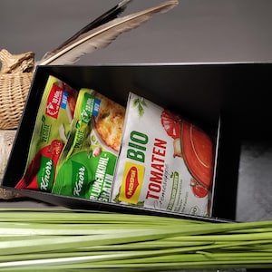 Puede incluir: Un recipiente negro que contiene varios paquetes de alimentos y una lata de sopa de tomate. Los paquetes están etiquetados como "Veggie" y "Knorr", mientras que la lata está etiquetada como "BIO TOMATEN Cremesuppe" con una ilustración de tomate.