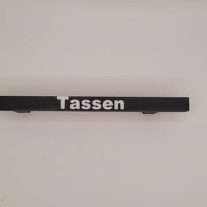 Op de afbeelding: Een zwart houten bord met het woord "Tassen" in wit gedrukt.