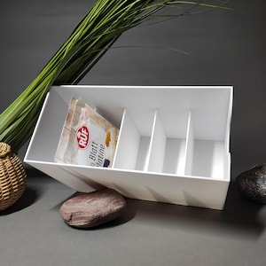 Op de afbeelding: Een witte rechthoekige organizer met drie interne verdelers, met een verpakking RUF Blatt Gelatine. De organizer is schuin geplaatst, met een minimalistisch ontwerp. Decoratieve elementen zijn een gevlochten mand, stenen en groen gras.