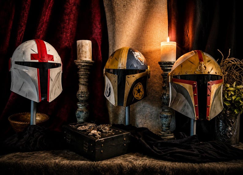 May include: Four Mandalorian helmets in various colors, including white with red accents, gold and black, and gold with red trim, are displayed on stands. A lit candle sits next to each helmet. A small treasure chest is in the foreground.
