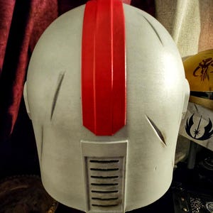 May include: A light gray helmet with a prominent red stripe down the center. The helmet has a rectangular vent and several etched lines. The design suggests a futuristic or science fiction theme.