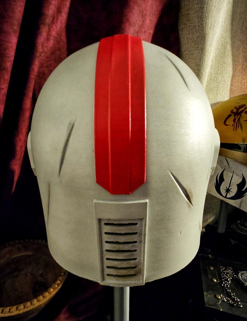 May include: A light gray helmet with a prominent red stripe down the center. The helmet has a rectangular vent and several etched lines. The design suggests a futuristic or science fiction theme.