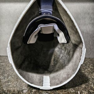 May include: Interior view of a white helmet with a black and white padded interior. The helmet's interior is dark gray, with a white padded chin strap and a black head support.