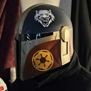 May include: A custom-painted helmet with a black and gold color scheme. The helmet features a white wolf emblem on the top and a gold and yellow emblem on the side. The helmet is designed with a visor and is likely for cosplay or display.