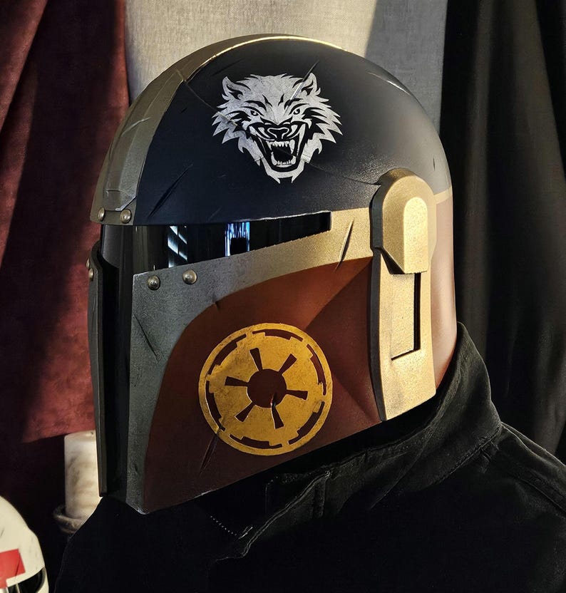 May include: A custom-painted helmet with a black and gold color scheme. The helmet features a white wolf emblem on the top and a gold and yellow emblem on the side. The helmet is designed with a visor and is likely for cosplay or display.