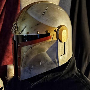 May include: A metallic silver and gold helmet with red accents. The helmet has a visor and is detailed with rivets and battle-worn scratches. The design is reminiscent of a Mandalorian helmet, suitable for cosplay or display.