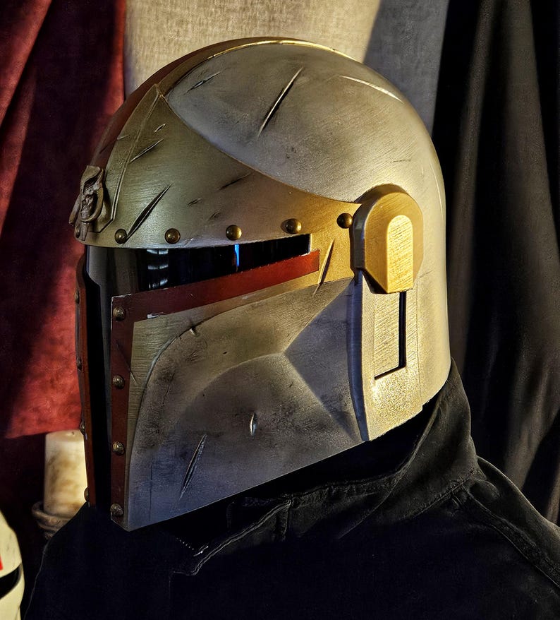 May include: A metallic silver and gold helmet with red accents. The helmet has a visor and is detailed with rivets and battle-worn scratches. The design is reminiscent of a Mandalorian helmet, suitable for cosplay or display.