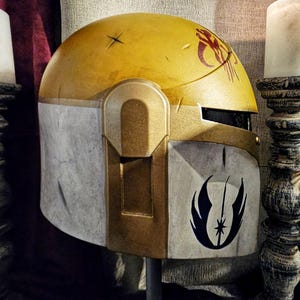 May include: A yellow and gray helmet with a gold visor. The helmet has a red symbol on the top and a black emblem on the side. The helmet is likely a costume piece or prop.