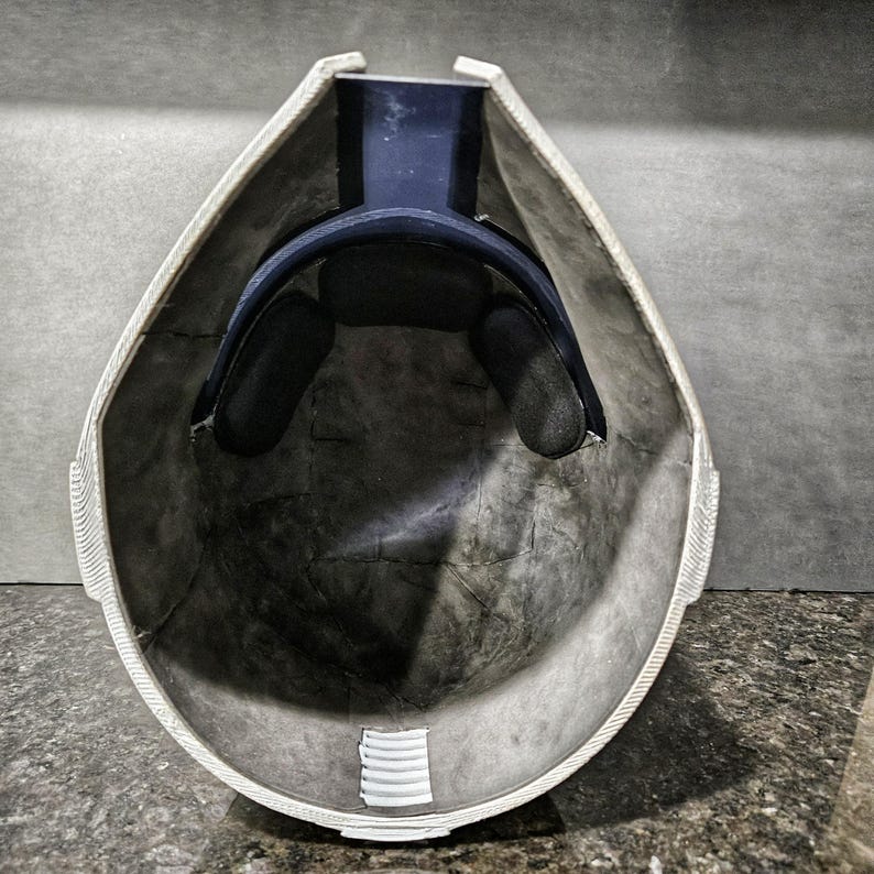 May include: An inside view of a white and gray helmet with a dark blue interior support and black padding. The helmet has a textured surface and a white trim around the edges. The interior is dark gray.