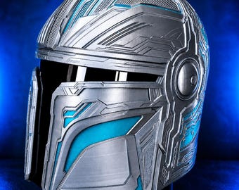 Könnte beinhalten: Ein silber-blauer Mandalorianer-Helm mit komplizierten geometrischen Mustern. Der Helm hat ein Visier und ist vor einem dunkelblauen Hintergrund platziert. Das Design umfasst detaillierte Paneele und Akzente.